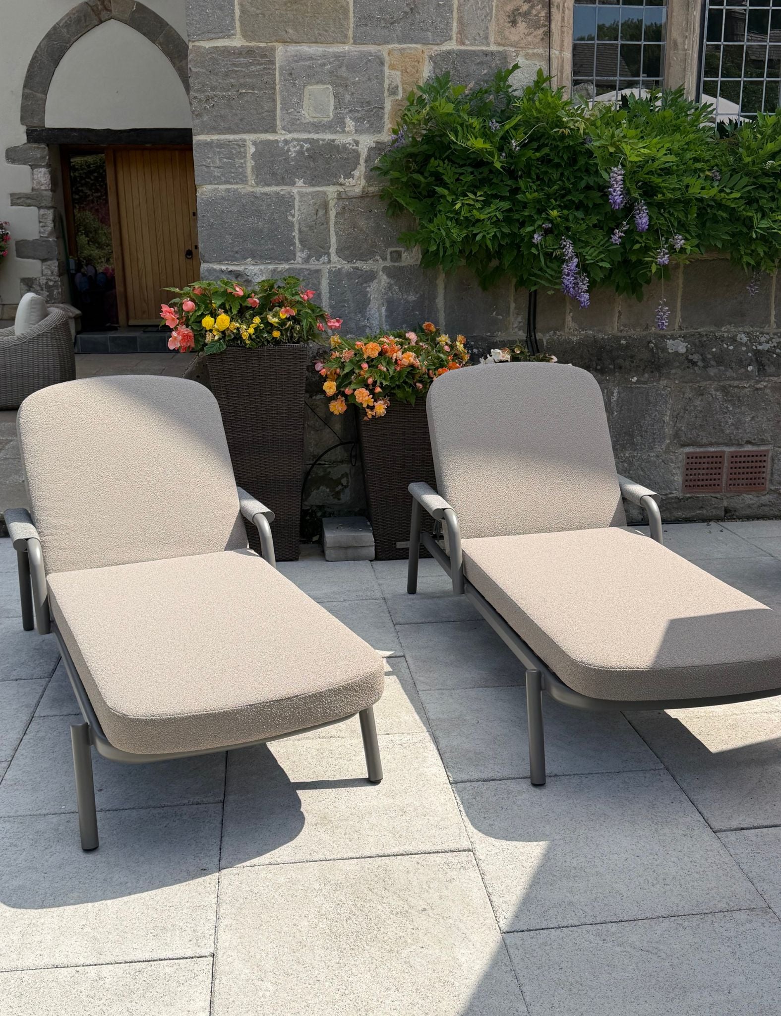 Image of Two outdoor chaise lounge chairs with beige cushions and dark metal frames, placed on a light stone patio in front of a stone building with potted flowers and greenery.
