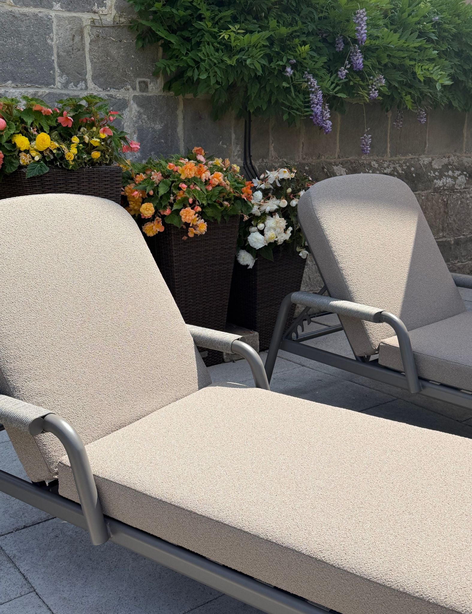 Image of A close-up of a beige outdoor chaise lounge chair with a silver metal frame, positioned on a stone patio next to another similar lounge chair, with potted flowers and a stone wall in the background.