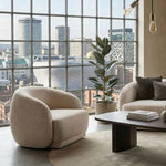 Modern living room with a large window view of a cityscape, featuring a beige armchair and a round coffee table.