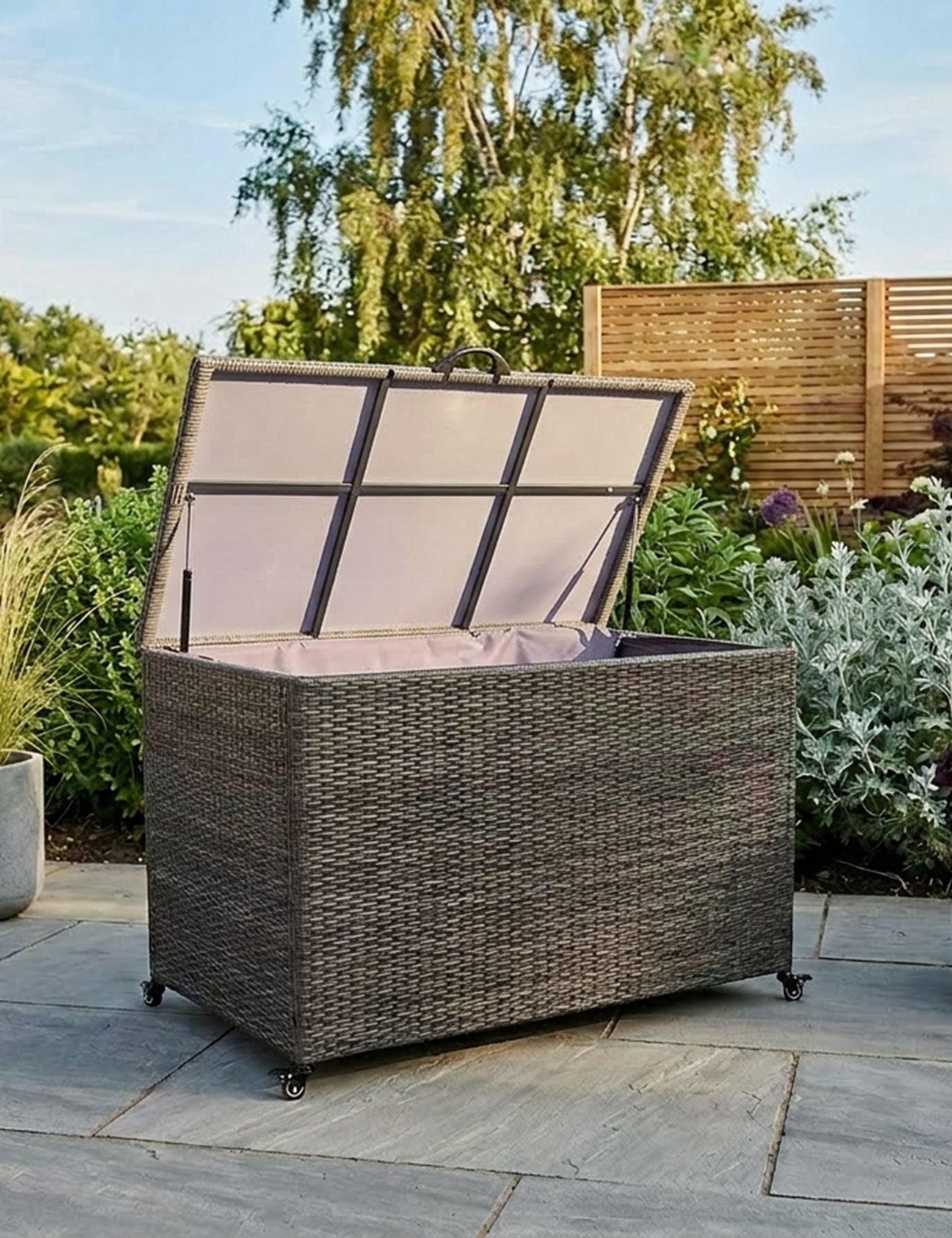 Image of A large Rattan storage chest with its lid open, set on a stone patio in a garden with plants and a wooden fence in the background.