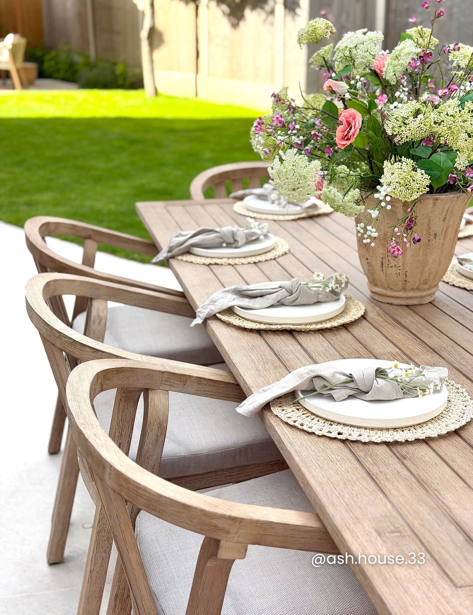 Outdoor dining table setup with wooden chairs, white plates, gray napkins, woven placemats, and a large floral centerpiece in a rustic bucket vase, set on a patio with a green lawn in the background.