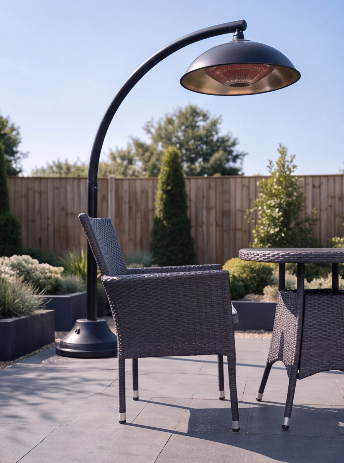 Image of A curved outdoor patio heater with a black metal frame and dome shade, positioned next to a dark gray wicker armchair on a paved patio with a wooden fence background.
