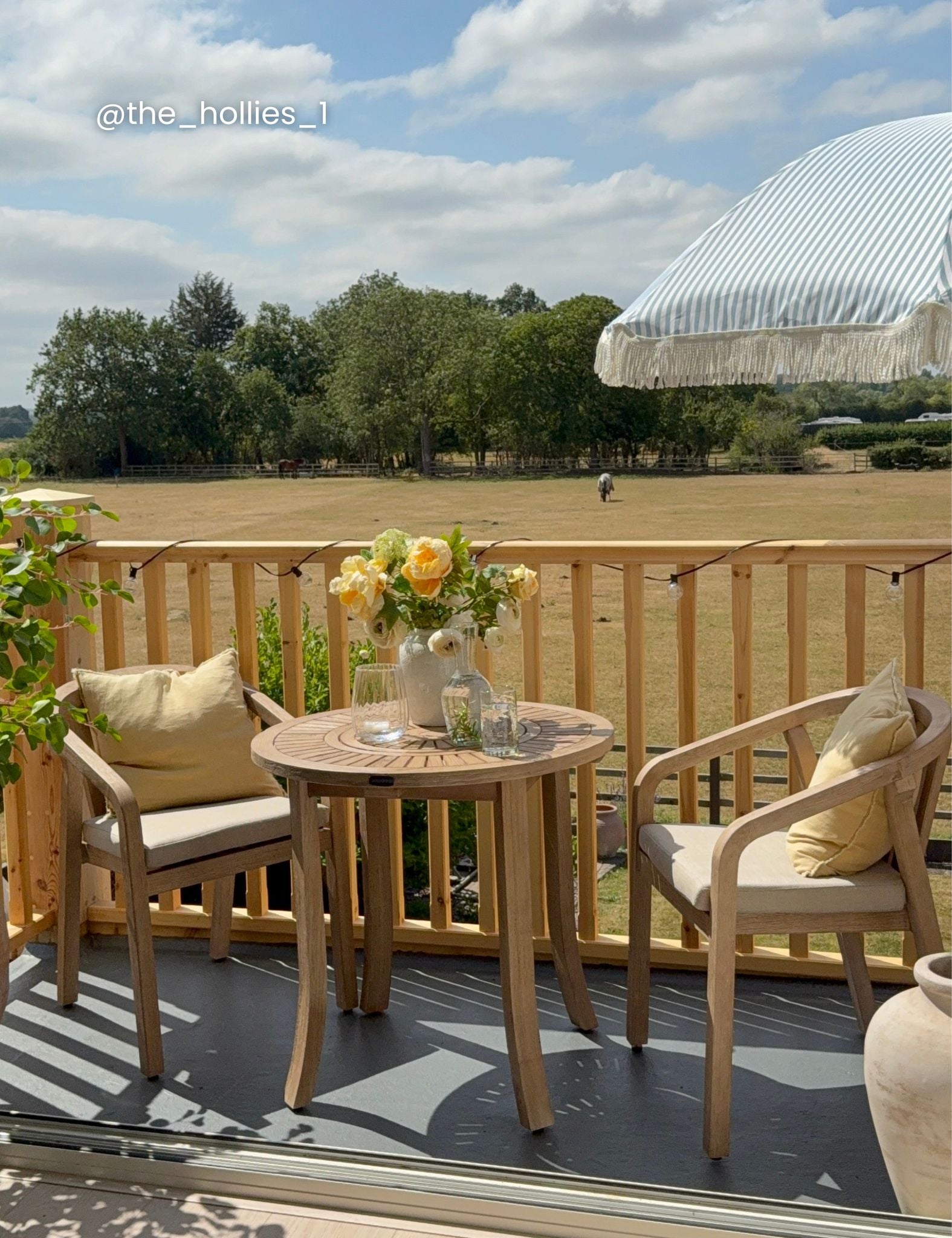 Image of Outdoor patio furniture set with two light wood armchairs, a matching round wooden table, and a vase of yellow roses on a deck overlooking a grassy field and trees under a blue sky.