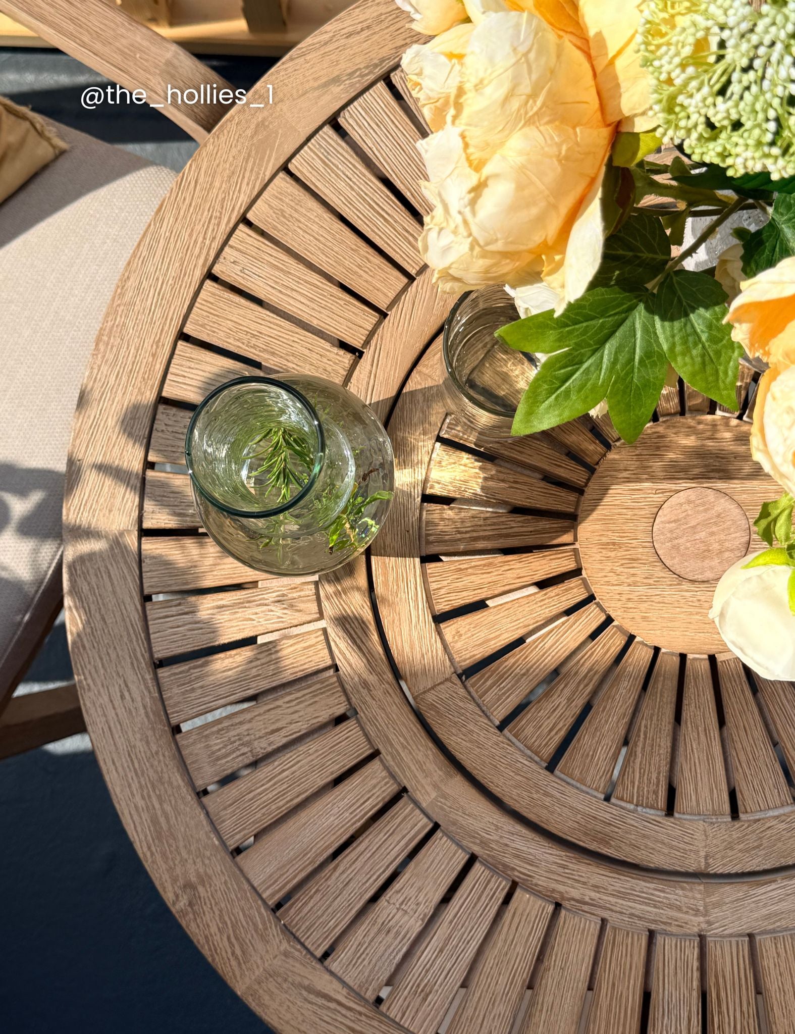 Image of A top-down view of a round wooden slat outdoor table with a glass vase and yellow flowers on a patio.