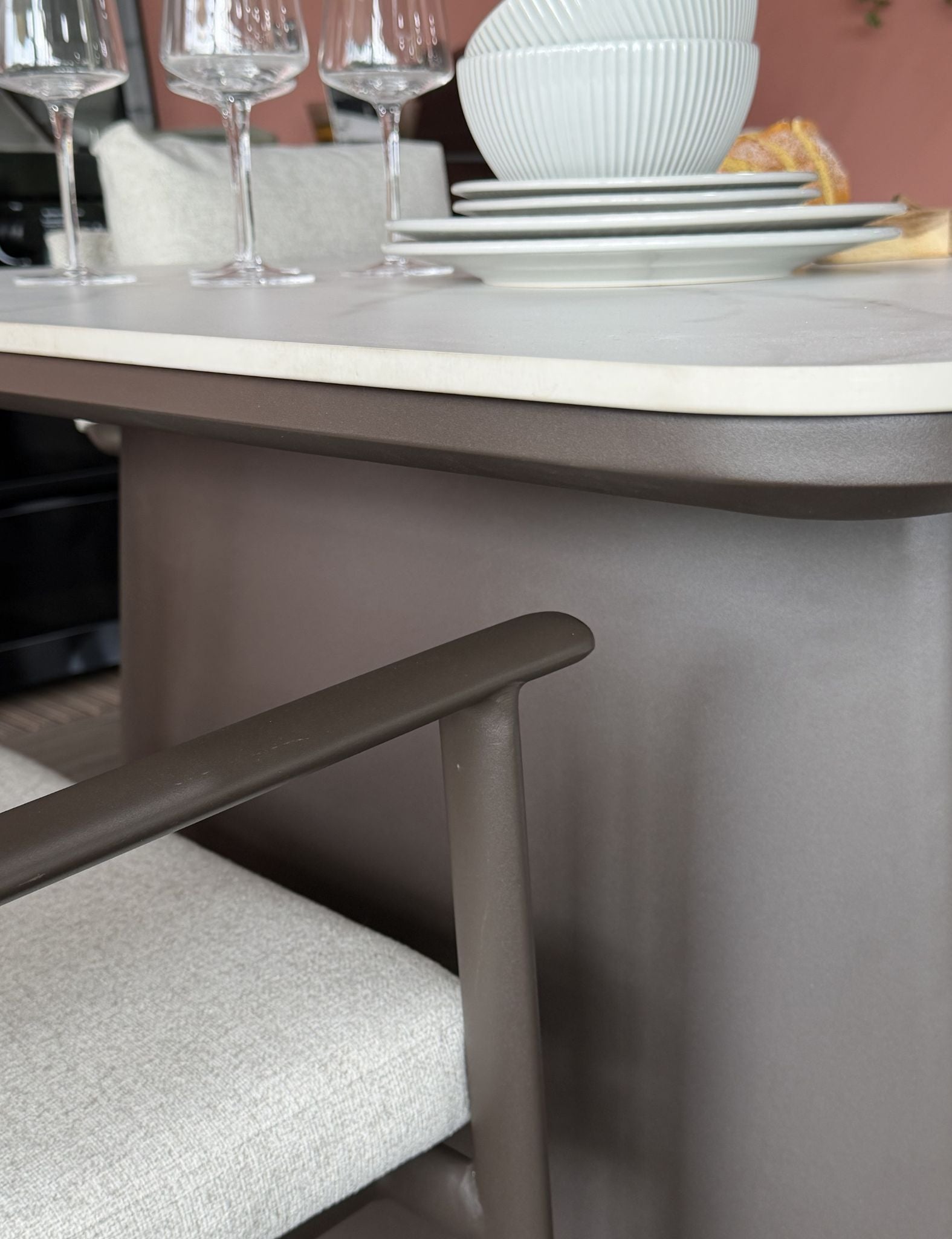 Image of Close-up of a dining table and chair in a real interior setting, with a white tabletop, taupe-gray table base, upholstered chair seat, wine glasses, stacked white plates, and a ribbed white bowl.