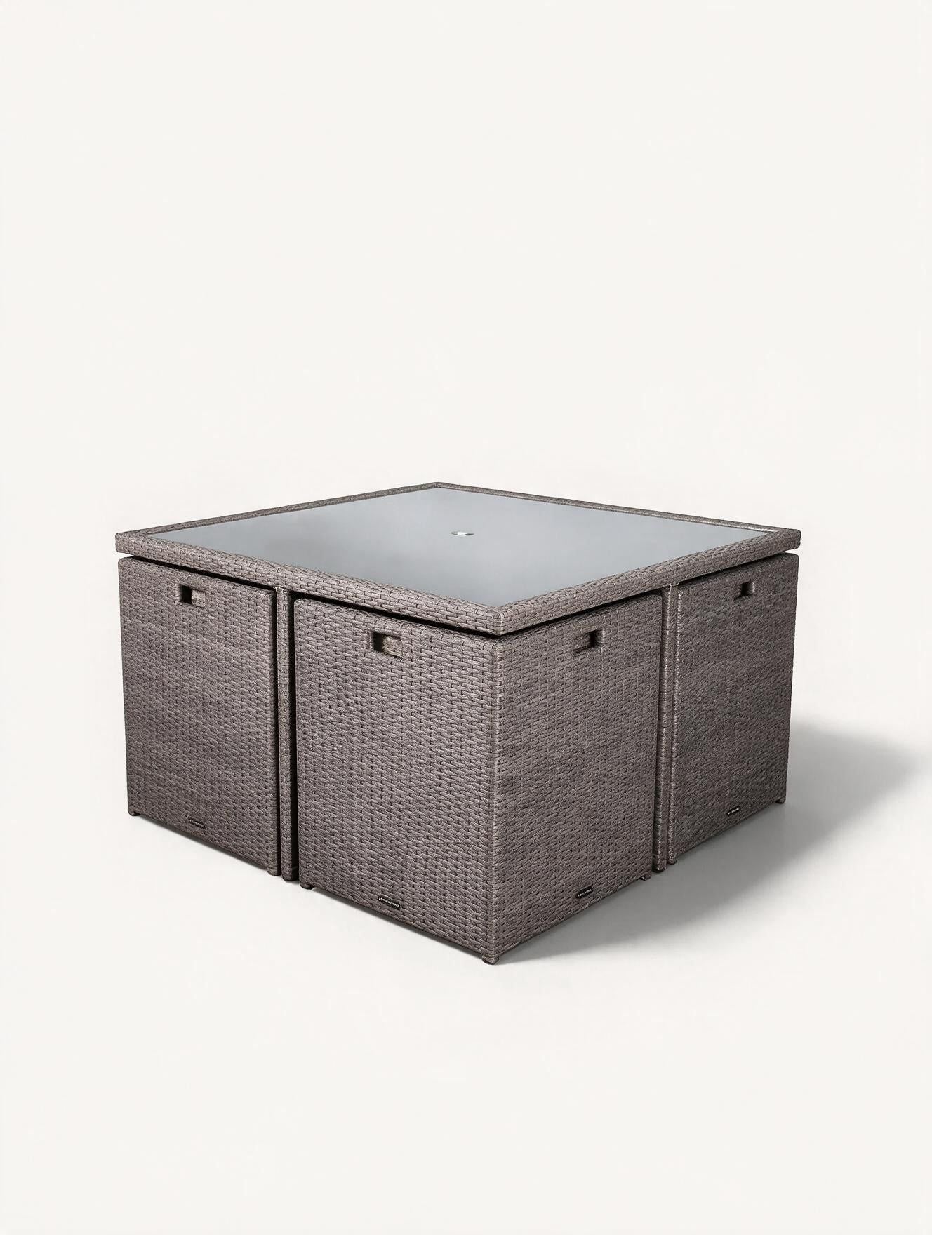 Image of A square outdoor patio table set with a Rattan grey body and a flat square glass top and chairs tucked underneath to form a compact cube, shown on a plain light background.