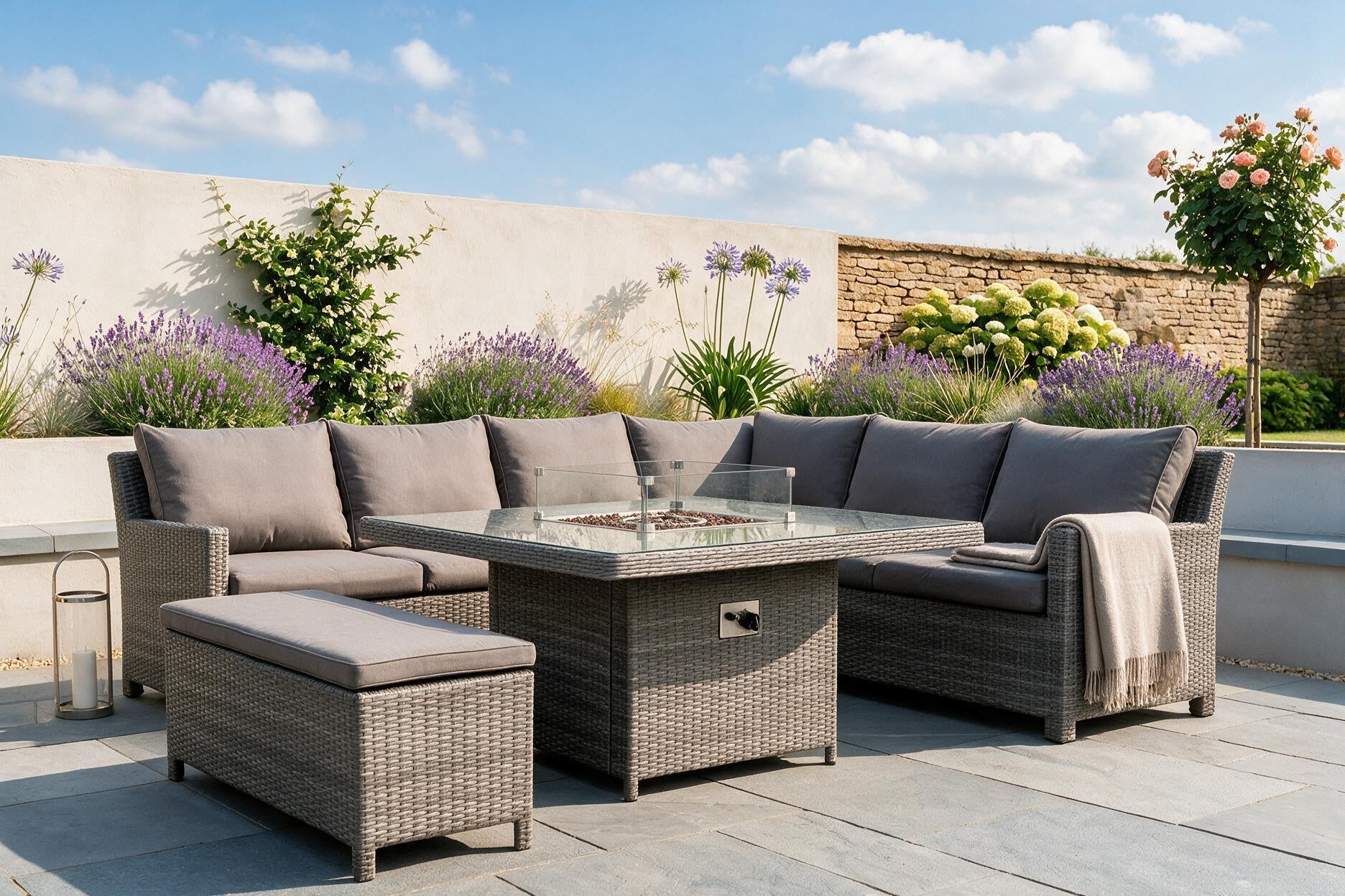A 5-piece outdoor patio furniture set featuring a gray wicker sectional sofa, matching ottoman, and a glass-top fire pit table, arranged on a stone patio with a garden background.