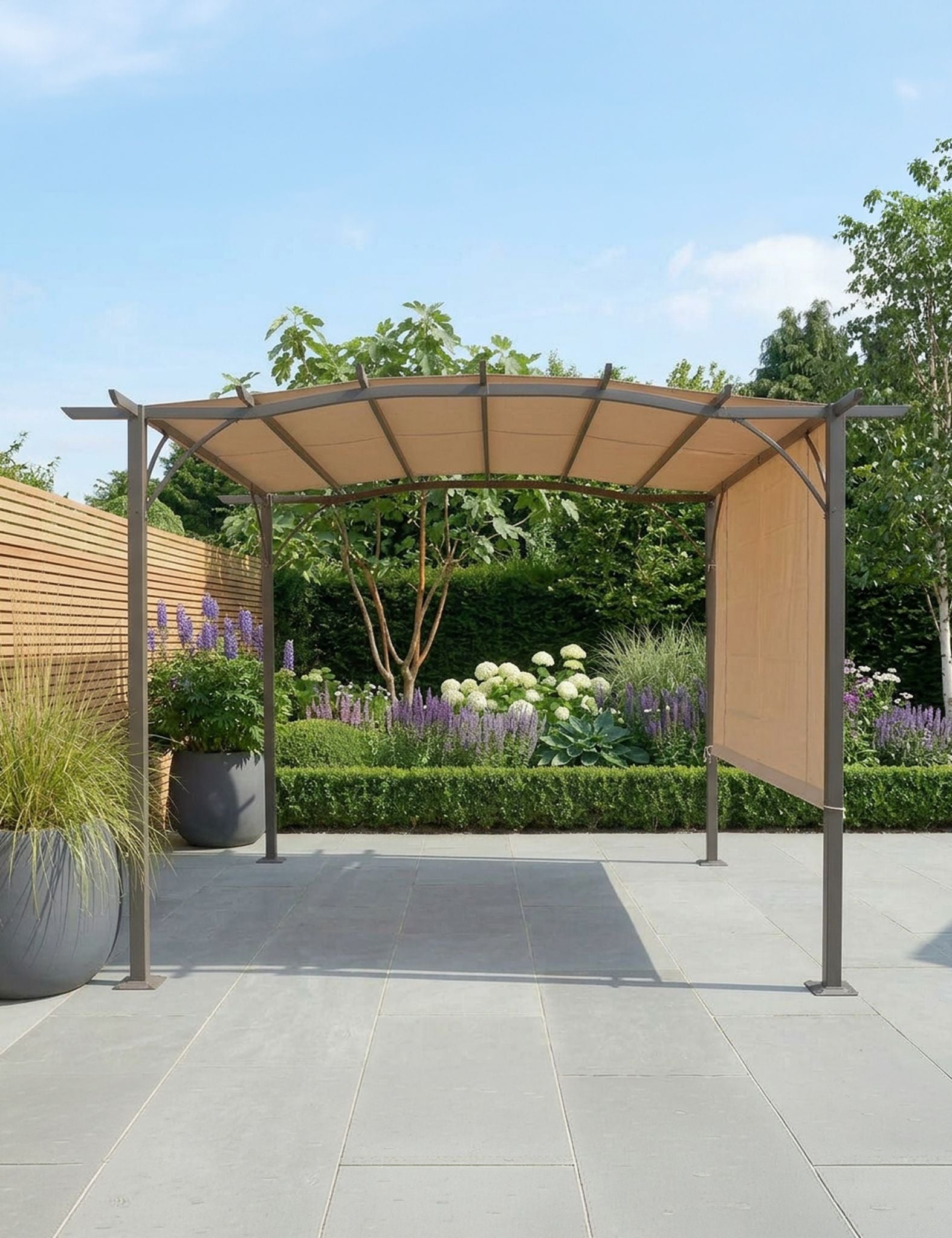 Image of A modern outdoor patio pergola with a curved beige canopy and dark metal frame, set on a paved patio with a landscaped garden background.