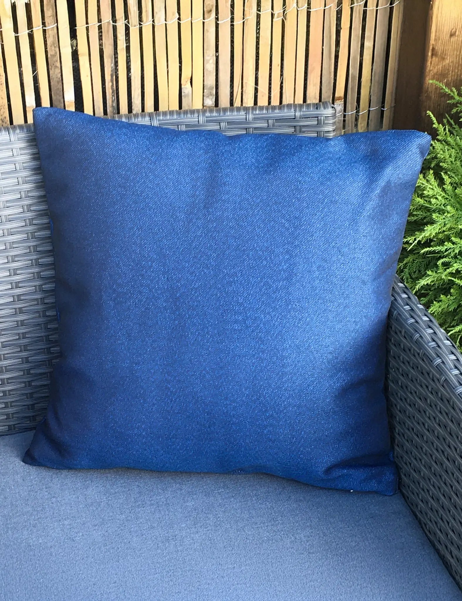 Image of Blue pillow on a wicker chair with a wooden fence and greenery in the background
