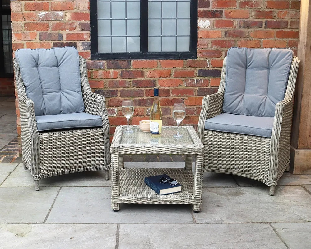 Image of Two wicker chairs with gray cushions and a glass table against a brick wall.