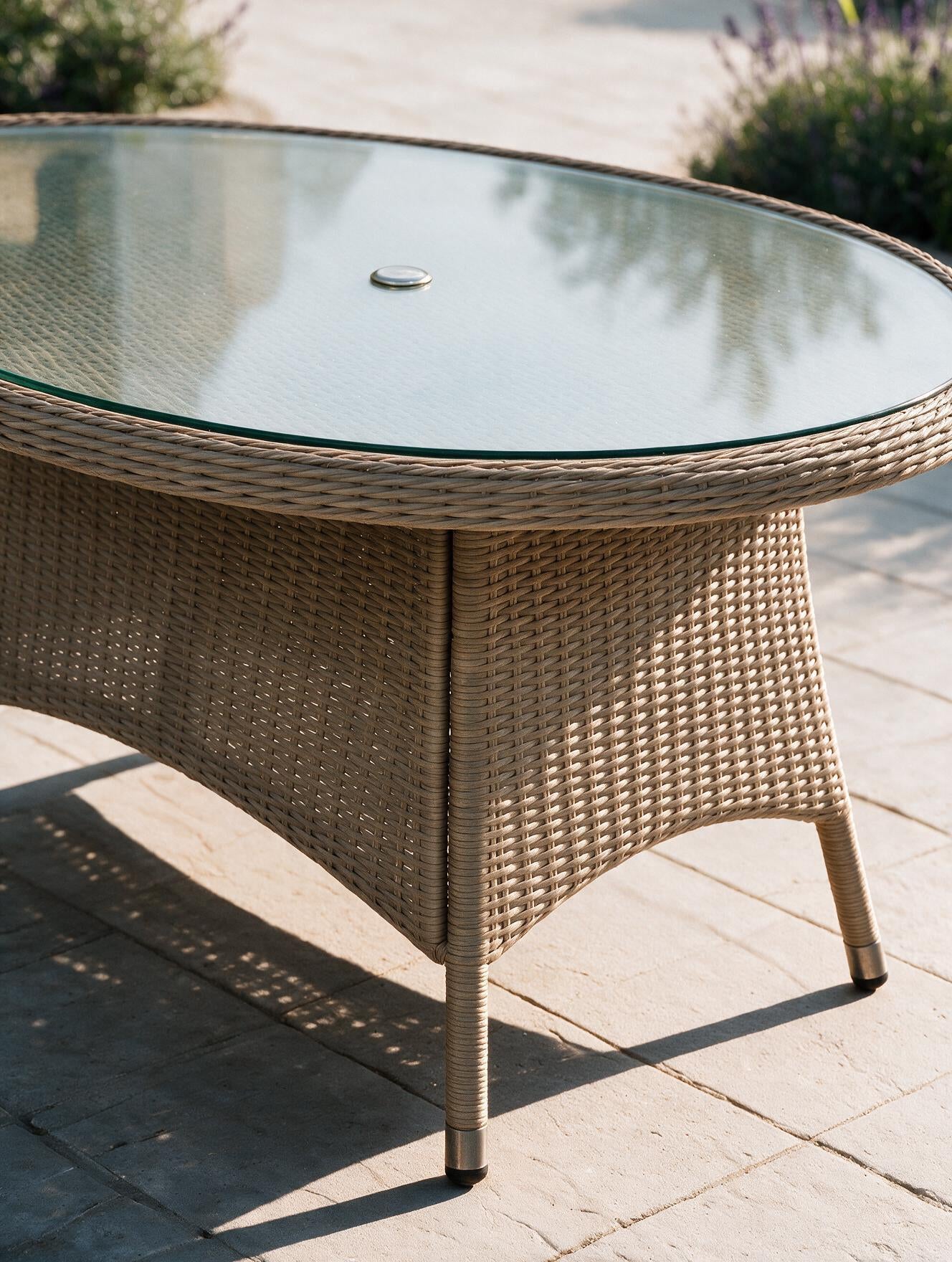 Image of An outdoor oval rattan dining table with a clear glass top, shown on a stone patio with garden plants in the background.