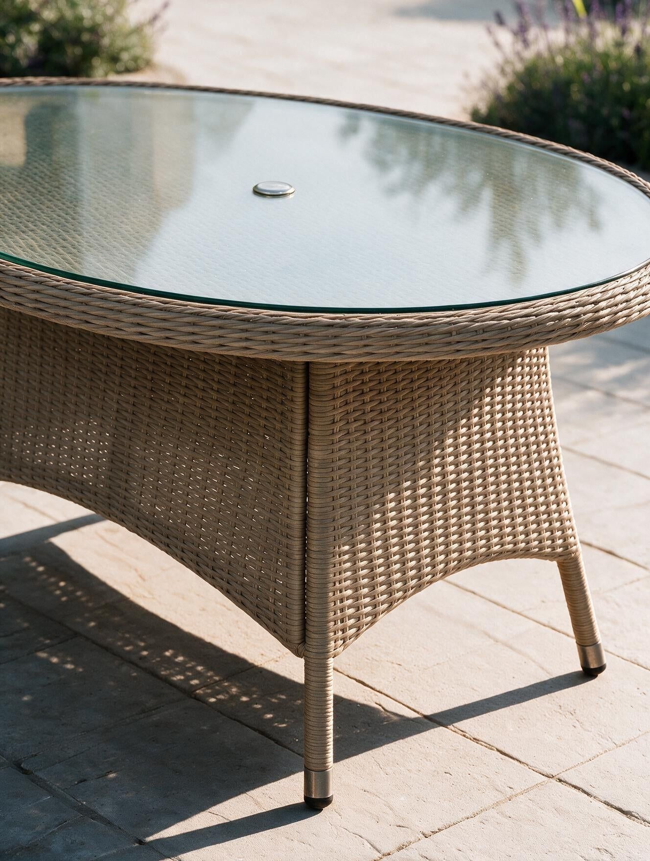 Image of An outdoor oval rattan dining table with a clear glass top, shown on a stone patio with garden plants in the background.