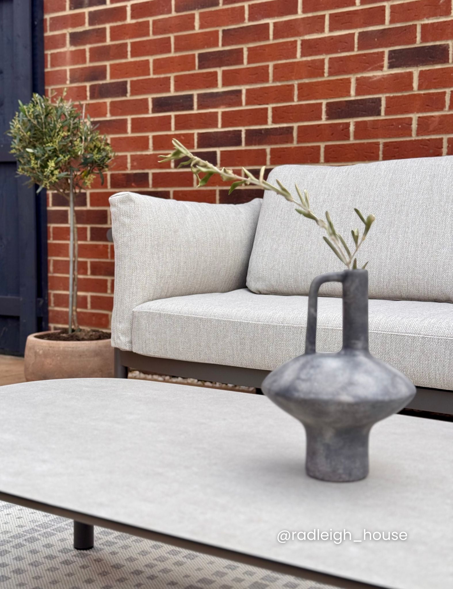 Image of Outdoor setting with a light gray sofa, white coffee table, and decorative vase against a brick wall.